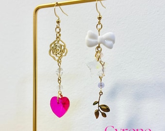 May include: A pair of asymmetrical dangle earrings. One earring features a gold-tone rose, clear beads, and a pink heart charm. The other has a white bow, star-shaped beads, and a gold-tone leaf. The word "Cyrene" is in pink.
