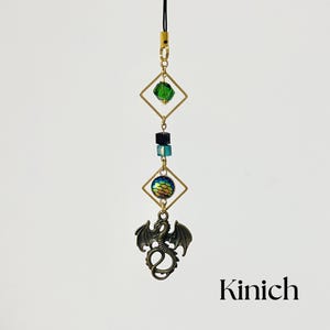 May include: A fantasy-themed charm with a bronze dragon pendant, iridescent orb, green and blue beads, and gold-coloured geometric shapes. The charm hangs from a black cord and includes the text "Kinich".