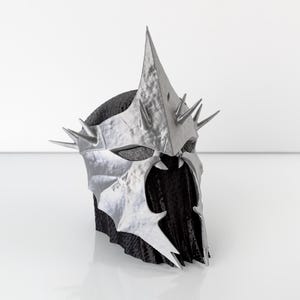 Peut inclure: Un casque argenté avec des pointes acérées et un design angulaire distinctif. Le casque repose sur une base sombre et texturée, sur fond blanc. Le design évoque des thèmes fantastiques ou médiévaux, avec des détails pointus.