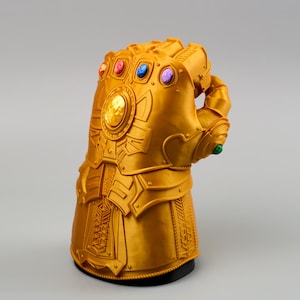 Op de afbeelding: Een gouden, gedetailleerde handschoen met zes kleurrijke edelstenen. De handschoen is een replica van de Infinity Gauntlet, een fictief item uit een stripboekenserie. De handschoen staat op een zwarte basis en heeft ingewikkelde ontwerpen.