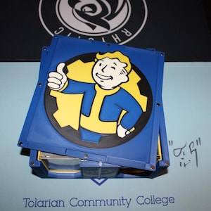 May include: A blue container with a yellow and black graphic of a character giving a thumbs up. The container is on a light blue surface with the text "Tolarian Community College" at the bottom. A black surface is in the background.