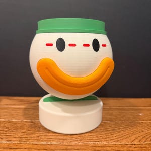 May include: A spherical white container with a green rim and a prominent orange curved mouth. The face features two black oval eyes and a series of red horizontal dashes above them. The object is supported by a cylindrical white base.