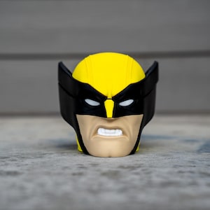 May include: A yellow and black Wolverine mask figurine with a stern expression. The mask features black eye coverings and ear-like protrusions, with a tan face and white teeth. The figurine is set against a gray background.