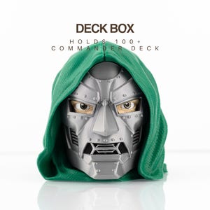 May include: A green and silver deck box shaped like a masked character head with a hood. The text DECK BOX and HOLDS 100+ COMMANDER DECK is printed above the object. The mask is metallic gray with visible rivets and a dark mouth grill.