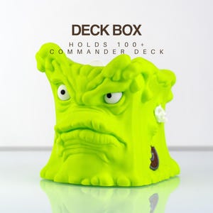 May include: A bright lime green deck box featuring a sculpted, grumpy monster face with white eyes. The text DECK BOX HOLDS 100+ COMMANDER DECK is printed above the container. The design has a textured, dripping appearance with a small circular detail.