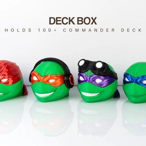 May include: Four green deck boxes shaped like Teenage Mutant Ninja Turtle heads, each with a different colored mask: red, orange, purple, and blue. The text "DECK BOX HOLDS 100+ COMMANDER DECK" is above the boxes.