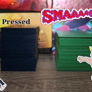 May include: Two stacks of playing cards, one navy blue and one green, are displayed. The text "Hand Pressed" is above the navy blue cards. The word "SMAAAASH!" is in pixelated letters above the green cards. Cartoon figures are on the image.