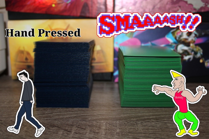 May include: Two stacks of playing cards, one navy blue and one green, are displayed. The text "Hand Pressed" is above the navy blue cards. The word "SMAAAASH!" is in pixelated letters above the green cards. Cartoon figures are on the image.