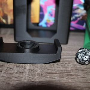 May include: A black 3D-printed card holder with a threaded center, designed to hold a deck of cards. A speckled black and white twenty-sided die sits next to the holder. Green and blue card decks are in the background.
