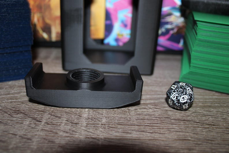 May include: A black 3D-printed card holder with a threaded center, designed to hold a deck of cards. A speckled black and white twenty-sided die sits next to the holder. Green and blue card decks are in the background.