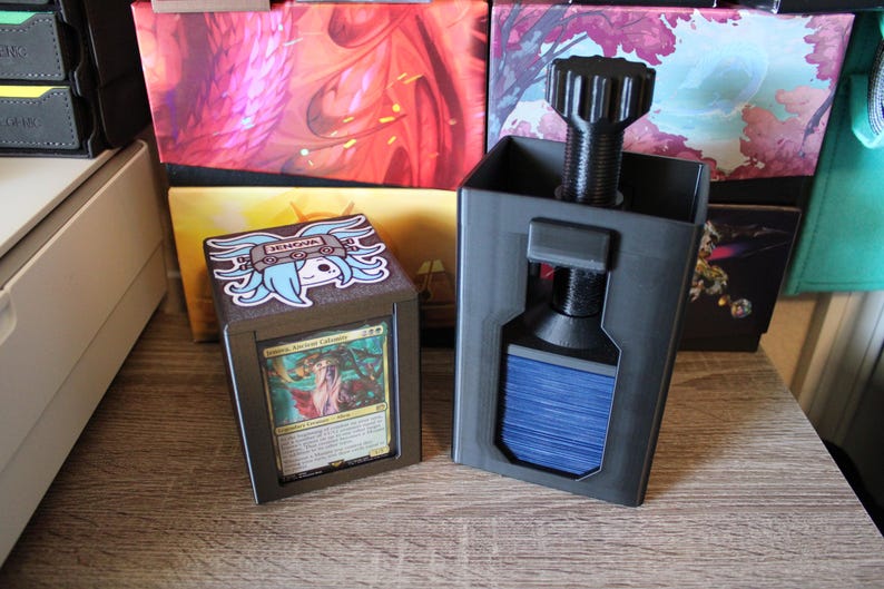 May include: Two black card storage boxes with a screw-on top. One box holds a card with a woman illustration. The other box has a sticker with a blue-haired anime character. The boxes are on a wooden surface.