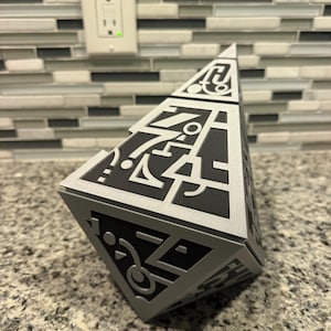 May include: A pyramid-shaped object featuring a matte black base with raised silver geometric patterns and abstract symbols on each triangular face. The structure has sharp edges and a pointed top, displaying a complex, layered design.