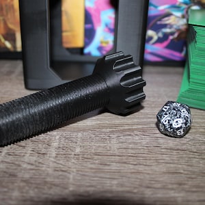 May include: Black, textured game accessory with a cylindrical body and a gear-like head. A speckled, multi-sided die is next to it. Stacks of blue and green cards are in the background, suggesting a tabletop gaming theme.
