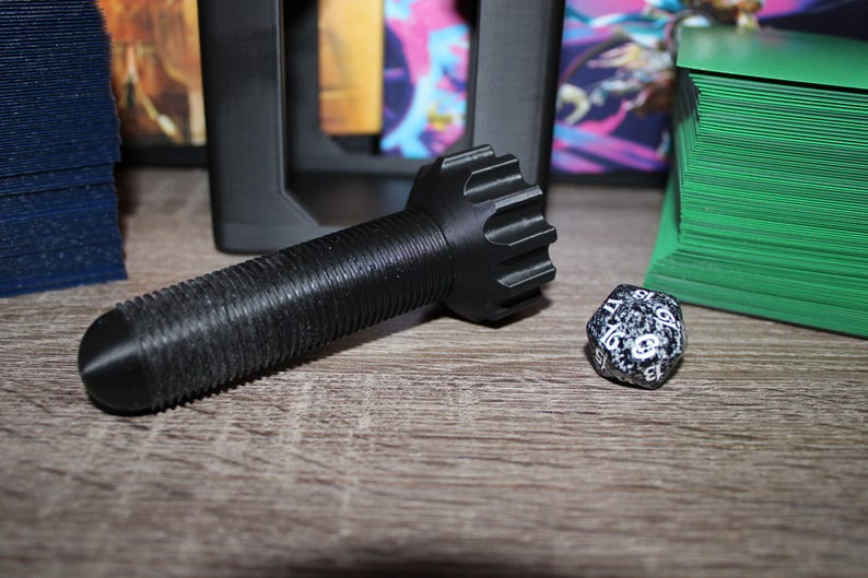 May include: Black, textured game accessory with a cylindrical body and a gear-like head. A speckled, multi-sided die is next to it. Stacks of blue and green cards are in the background, suggesting a tabletop gaming theme.
