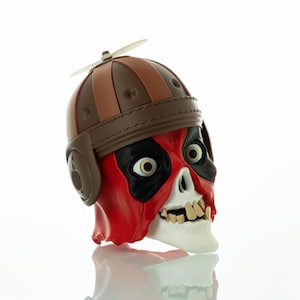 Könnte beinhalten: Eine neuartige Totenkopf-Figur mit einem braunen und beigefarbenen Fliegerhelm und einem kleinen Propeller. Der Totenkopf hat eine rot-schwarze Maske, weiße Zähne und große Augen.