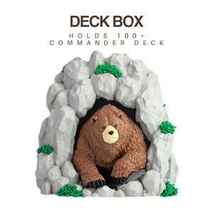 May include: A grey rock-shaped deck box with the text DECK BOX HOLDS 100+ COMMANDER DECK. The container features a brown bear emerging from a dark cave entrance, accented with small green tufts of vegetation on the exterior rock surface.