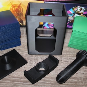 May include: A collection of black 3D-printed card game accessories, including a card holder, deck boxes, and a card shuffler. Stacks of blue and green cards are also present. The items are arranged on a wooden surface.