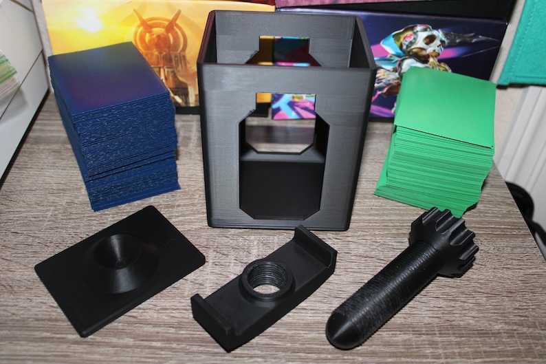 May include: A collection of black 3D-printed card game accessories, including a card holder, deck boxes, and a card shuffler. Stacks of blue and green cards are also present. The items are arranged on a wooden surface.