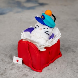 May include: A novelty figurine of a white cat wearing a blue and orange hat, sitting in a red postbox filled with white paper. A small white envelope with a red dot is placed next to the postbox. The cat has black whiskers and purple accents.