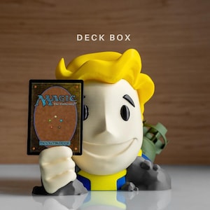 May include: A 3D-printed deck box shaped like a cartoon character with bright yellow hair, a cream-colored face, and a blue and yellow outfit. The character holds a Magic: The Gathering card in its hand. The text DECK BOX is printed above the figure.