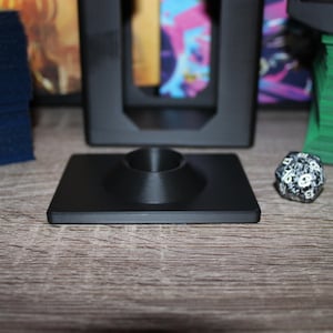 May include: A black square gaming accessory with a central conical support, likely for holding a game piece. A multi-sided die and other game accessories are visible in the background. The item is on a wooden surface.