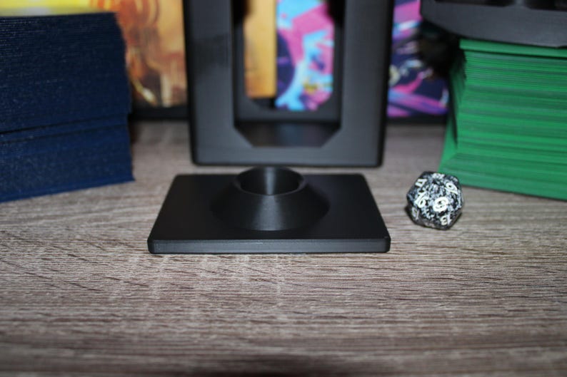 May include: A black square gaming accessory with a central conical support, likely for holding a game piece. A multi-sided die and other game accessories are visible in the background. The item is on a wooden surface.