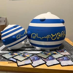 May include: Two spherical containers with blue and white stripes and rounded blue accents. The larger container has the word "Blupal" in yellow script. Several trading cards are displayed in front of the containers.