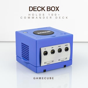 May include: A blue deck box designed to resemble a game console, featuring a silver front panel with four controller ports and a circular black top lid. The text DECK BOX, HOLDS 100+ COMMANDER DECK, and GAMECUBE are printed on the image.