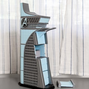 Puede incluir: Modelo de la Torre de los Vengadores en azul claro y gris, con el logotipo "A" de los Vengadores. El modelo tiene una base rectangular y una fachada detallada con una sección curva y una pequeña plataforma separada.