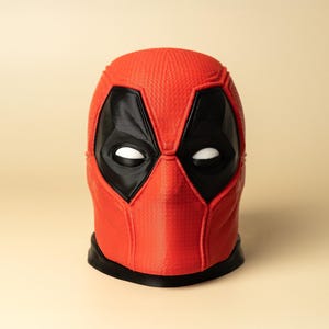 May include: A red and black Deadpool mask with white eyes. The mask is made of a textured material and has a detailed design.