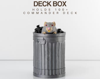 Puede incluir: Una caja de barajas gris con forma de cubo de basura, con una rata de dibujos animados asomando por la parte superior. El texto "DECK BOX" y "HOLDS 100+ COMMANDER DECK" están impresos encima de la caja. El texto "TRASH RAT" está impreso debajo.