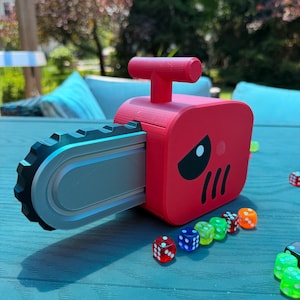 May include: A red chainsaw-shaped object with a grey blade and a black gear-like edge. The red body features a black eye graphic and three black vertical lines. Several small, translucent, multi-colored dice are scattered in front of the object.