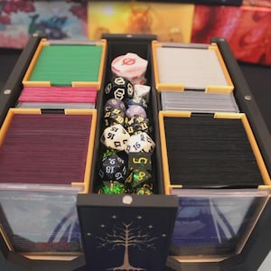 May include: A black and yellow tabletop gaming organiser, filled with stacks of cards in various colours, including green, pink, and black. Dice of different sizes and colours are in the centre. The organiser has a tree design on the front.