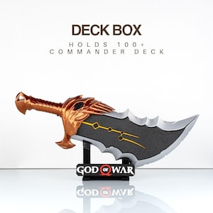 Op de afbeelding: Een deck box in de vorm van een zwaard met een metalen koperkleurig handvat en een donkergrijs blad met gele geometrische lijnen. De tekst DECK BOX HOLDS 100+ COMMANDER DECK staat erboven en een zwarte standaard met GOD OF WAR eronder.