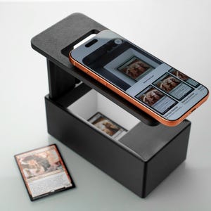 May include: A black card grading device with a smartphone resting on top. The phone displays images of trading cards. A trading card is visible on the surface next to the device. The device has a compartment to hold cards.