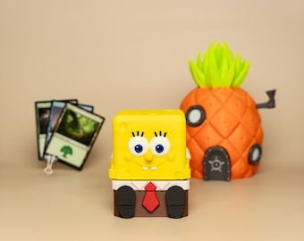 Puede incluir: Un personaje de dibujos animados amarillo y marrón con corbata, que se parece a Bob Esponja, se sienta frente a una casa con forma de piña. El personaje es una caja de almacenamiento para cartas de juego, con cuatro cartas visibles detrás de él.