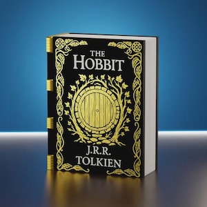 May include: A hardback copy of "The Hobbit" by J.R.R. Tolkien. The book has a black cover with gold detailing, including the title, author's name, and a circular illustration of a door. The pages are white, and the spine has gold accents.