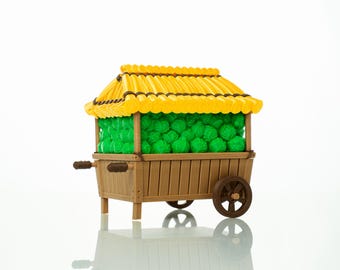 May include: A miniature wooden market cart with a yellow roof and brown accents. The cart is filled with green spheres, possibly representing produce. The cart has wheels and handles, suggesting it is mobile.