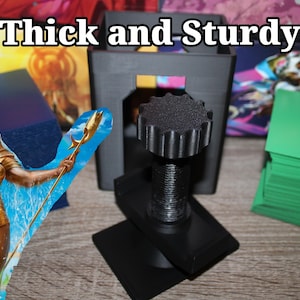 May include: A black, 3D-printed card holder with a threaded central column and a large, textured knob. The text "Thick and Sturdy" is at the top. A stack of green cards and a fantasy art card are also visible.