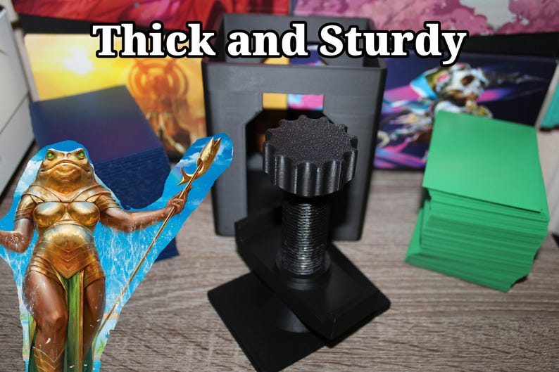 May include: A black, 3D-printed card holder with a threaded central column and a large, textured knob. The text "Thick and Sturdy" is at the top. A stack of green cards and a fantasy art card are also visible.