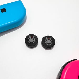 May include: Two black and grey Overwatch D.Va bunny themed joystick caps for a Nintendo Switch controller.