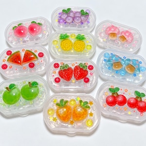 May include: A collection of ten clear, oval-shaped plastic containers, each filled with clear slime and decorated with a pair of colourful, three-dimensional charms including peaches, grapes, mushrooms, pineapples, watermelon slices, strawberries, apples, and cherries.