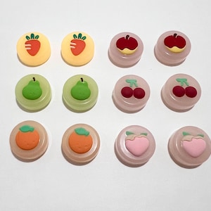May include: Twelve round, colourful, fruit-themed, plastic buttons. The buttons feature images of strawberries, pears, cherries, apples, peaches, and oranges.
