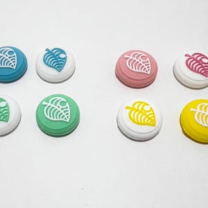 May include: Eight different coloured silicone button caps for a video game controller. The caps feature a white leaf design with a small circle in the centre.