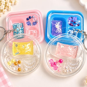 Peut inclure: Quatre porte-clés en plastique transparent avec des compartiments contenant des bonbons en résine colorés et des perles assorties. Deux boîtiers sont carrés avec des cadres rose et bleu, et deux sont circulaires. Ils contiennent des bonbons jaunes, roses et violets.