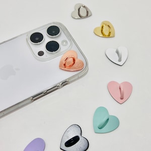 Peut inclure: Assortiment de supports d'anneau de téléphone en forme de cœur dans différentes couleurs, notamment or rose, argent, noir, blanc, rose, bleu clair et violet. Les anneaux sont conçus pour être fixés à l'arrière d'une coque de téléphone pour une prise en main sûre.