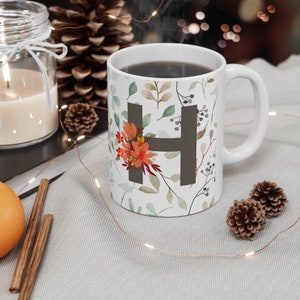 Puede incluir: Taza de cerámica blanca con un patrón floral y la letra "H" en gris. La taza está llena de café y está sobre una superficie blanca con una cadena de luces.