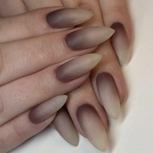Puede incluir: Primer plano de una mano con uñas largas en forma de almendra. Las uñas presentan un diseño degradado, que va del marrón oscuro en la base al beige claro en las puntas. Las uñas son lisas y están bien cuidadas.