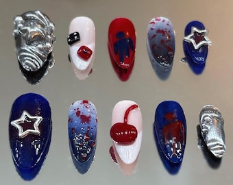 May include: A collection of ten artificial nails with various designs. The nails feature a mix of colors including red, blue, white, and silver. Some nails have star, dice, and cherry designs. Others have abstract patterns and small silver sculptures.