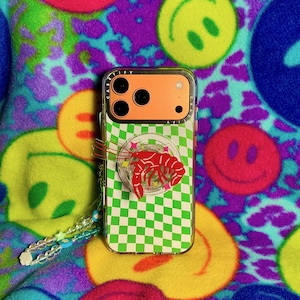 May include: A clear phone case with a green and white checkered pattern and a red lobster pop socket. The phone case has an orange back and is on a colorful smiley face background.